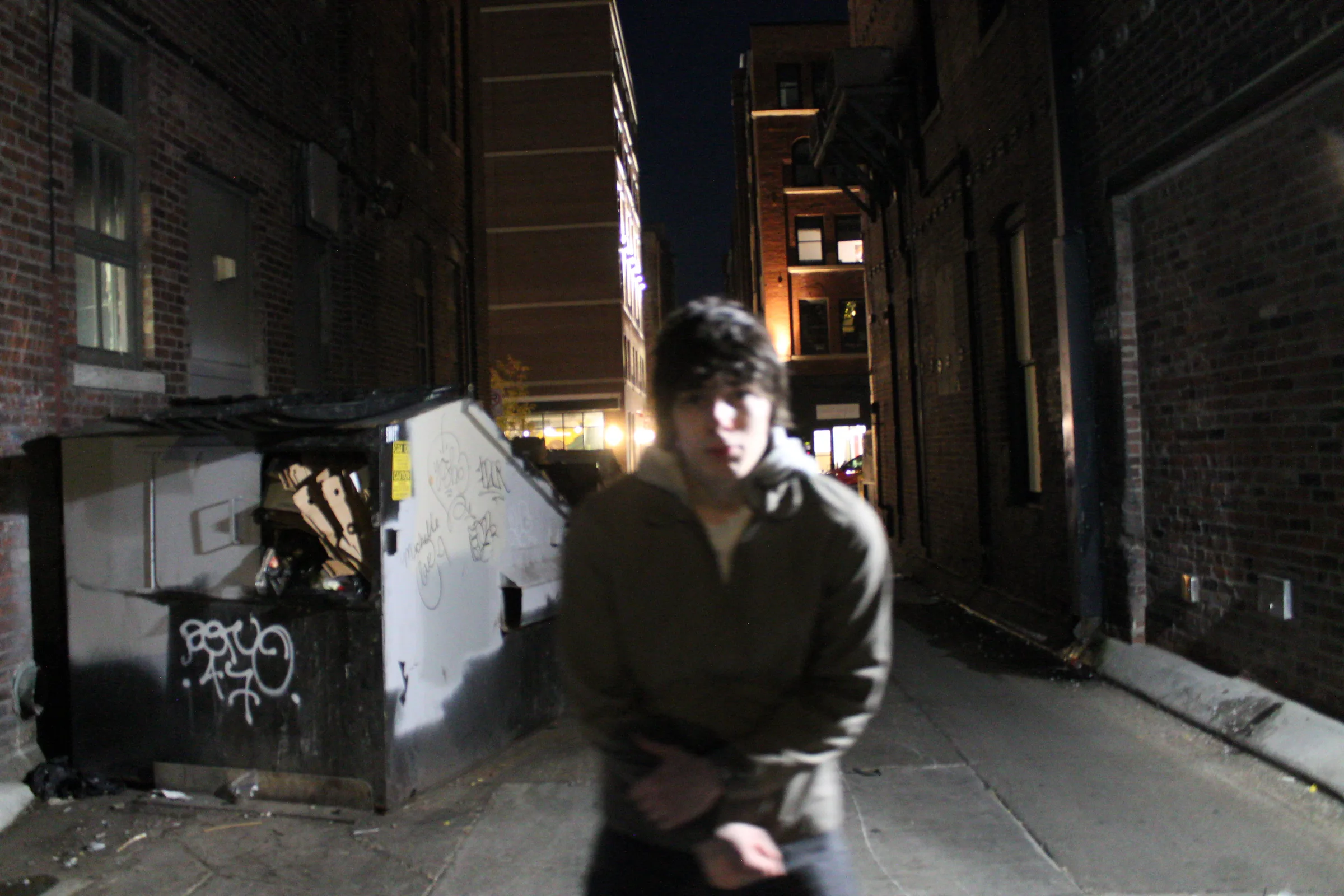 A person in a khaki jacket standing lit by flash in a narrow brick alley at night, a graffitied dumpster to one side and lit storefronts far behind.