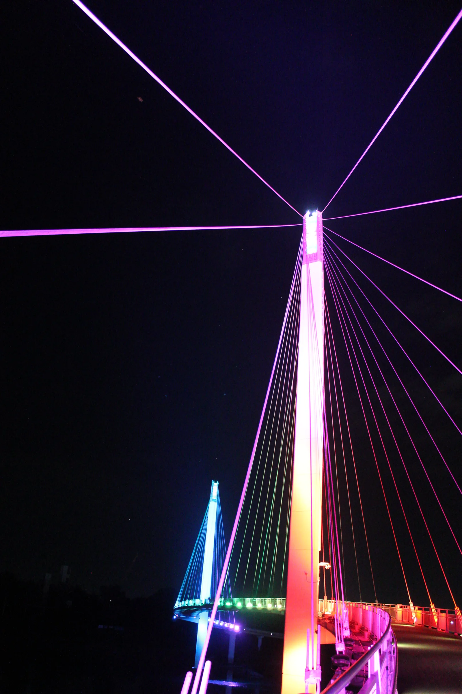 Looking up the tower of a lit pedestrian suspension bridge at night, its cables fanning out in magenta and orange, a second tower glowing blue-green beyond.