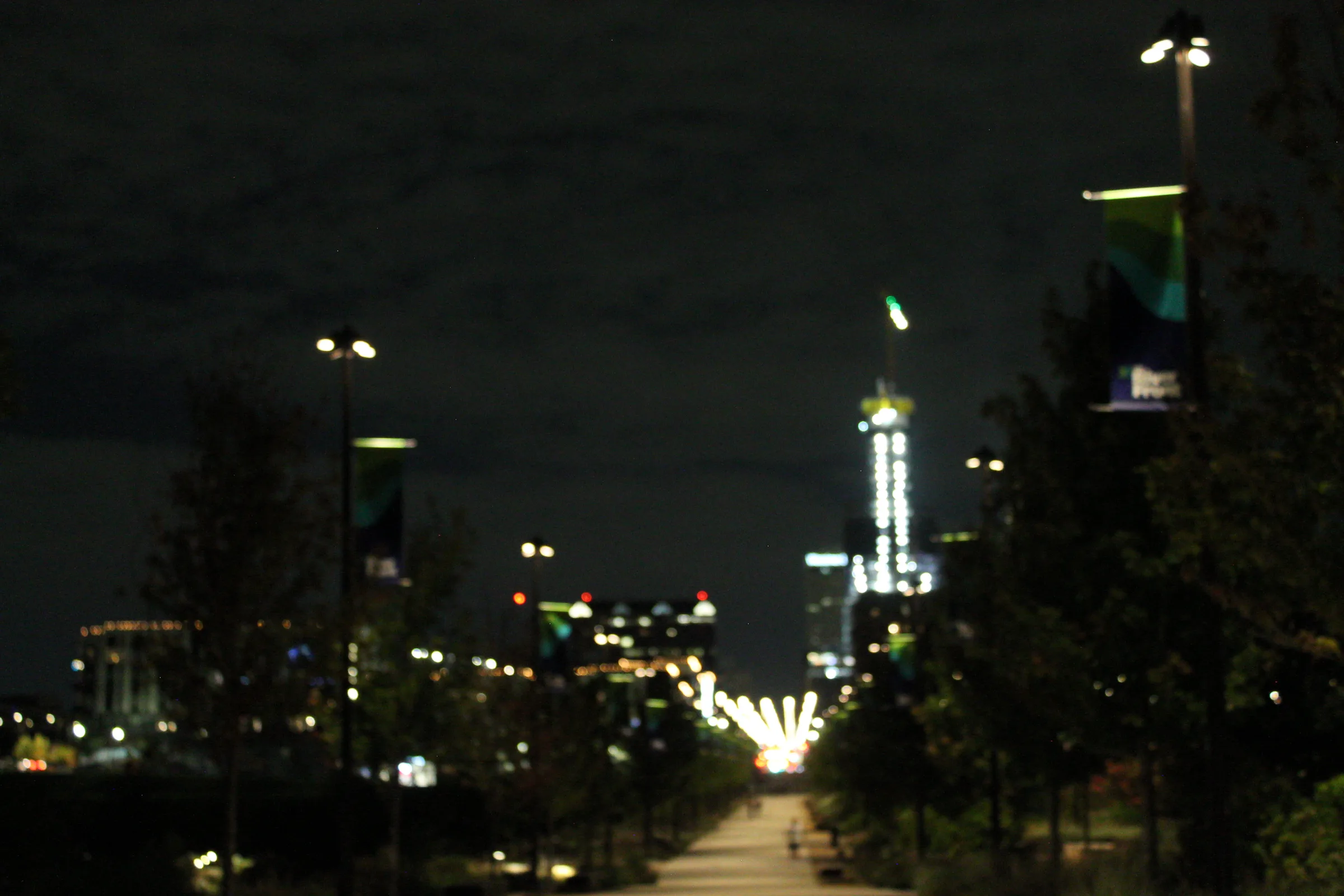 A tree-lined promenade at night, thrown out of focus, receding toward a brightly lit skyscraper with street lamps trailing away on either side.