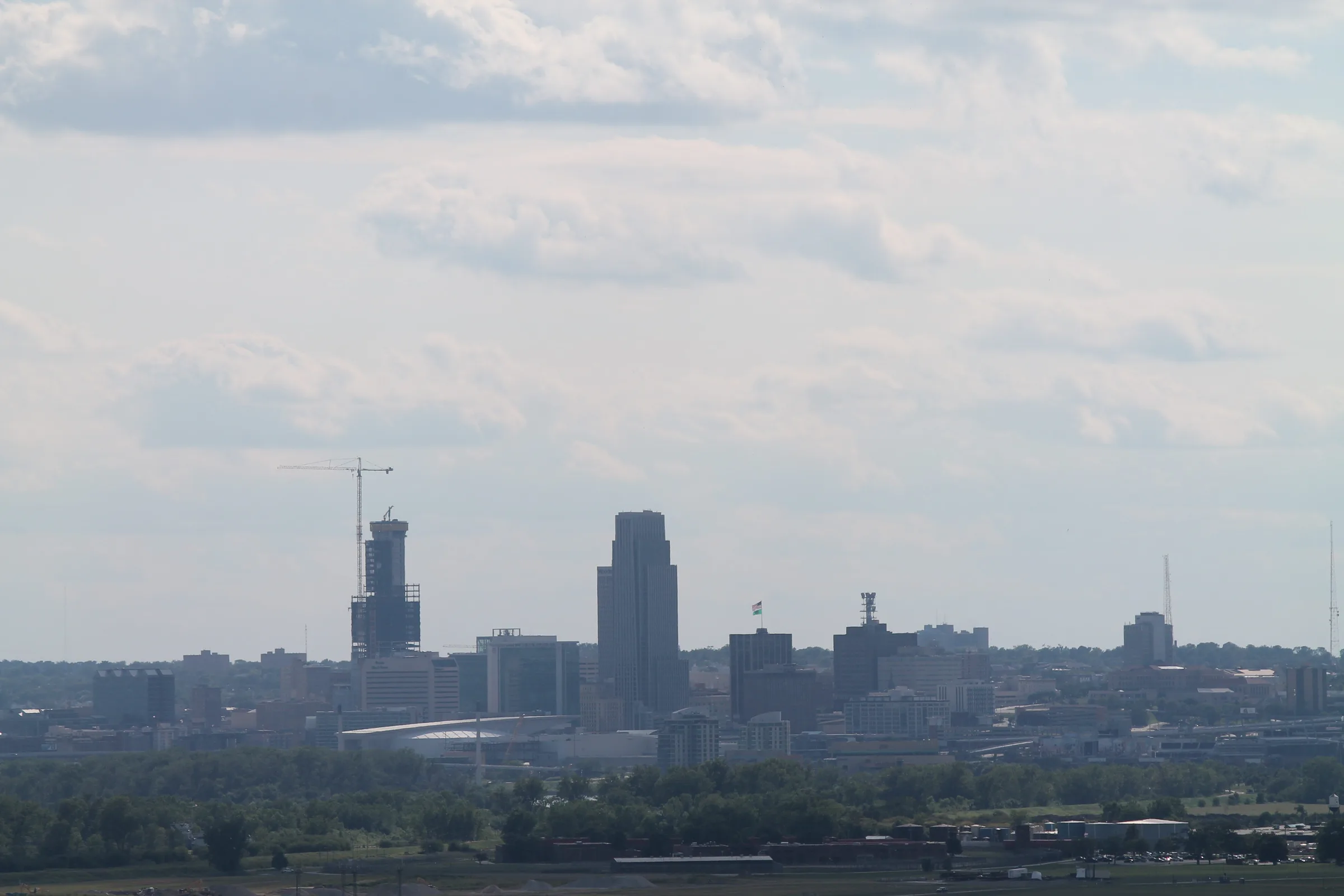 The Omaha skyline seen from a distance across low green riverbank, a construction crane beside a half-built tower under flat summer cloud.